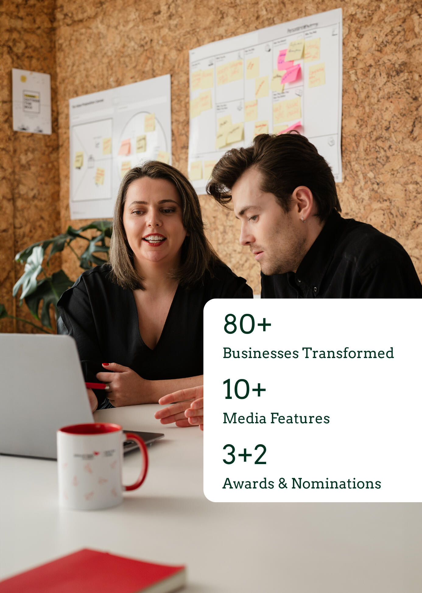 Kitti Majoran is working with a client, sitting at a desk in an office and looking at a laptop. There is also a graphic in the bottom right corner of the photo. On the graphic, the following is written: 70+ businesses transformed, 10+ media features, 3+2 a