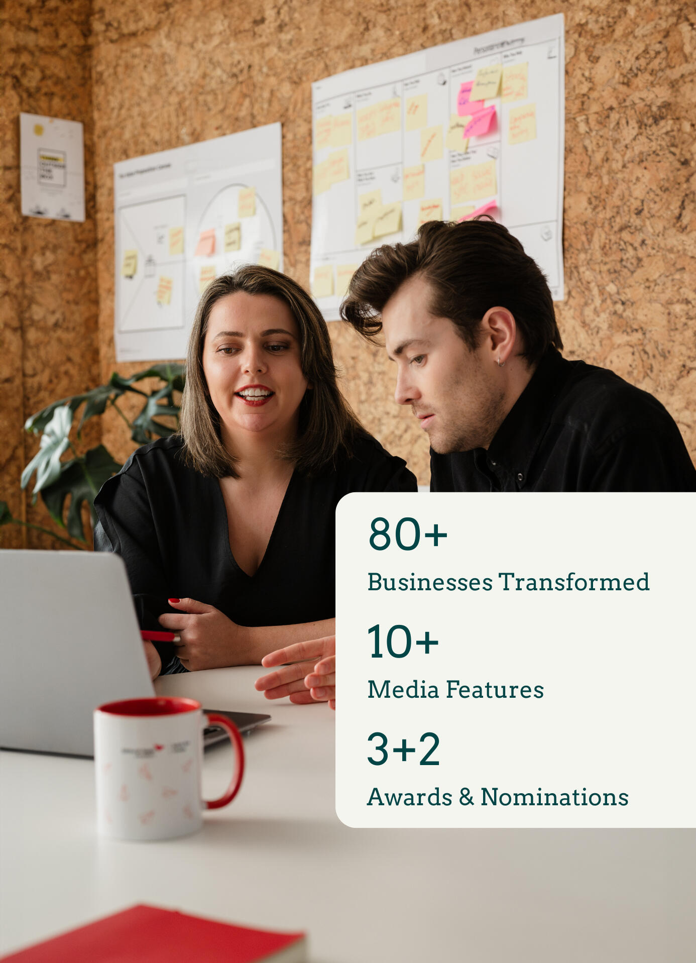 Kitti Majoran is working with a client, sitting at a desk in an office and looking at a laptop. There is also a graphic in the bottom right corner of the photo. On the graphic, the following is written: 80+ businesses transformed, 10+ media features, 3+2 a
