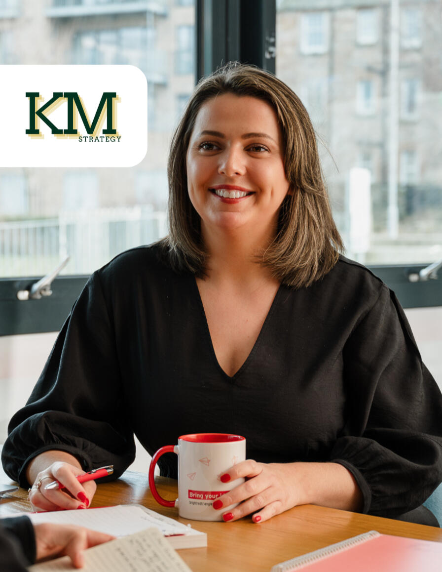 Kitti Majoran is sitting at a desk with a cup of coffee and a pen in her hand, smiling. The KM Strategy logo is also in the top left corner of the photo.